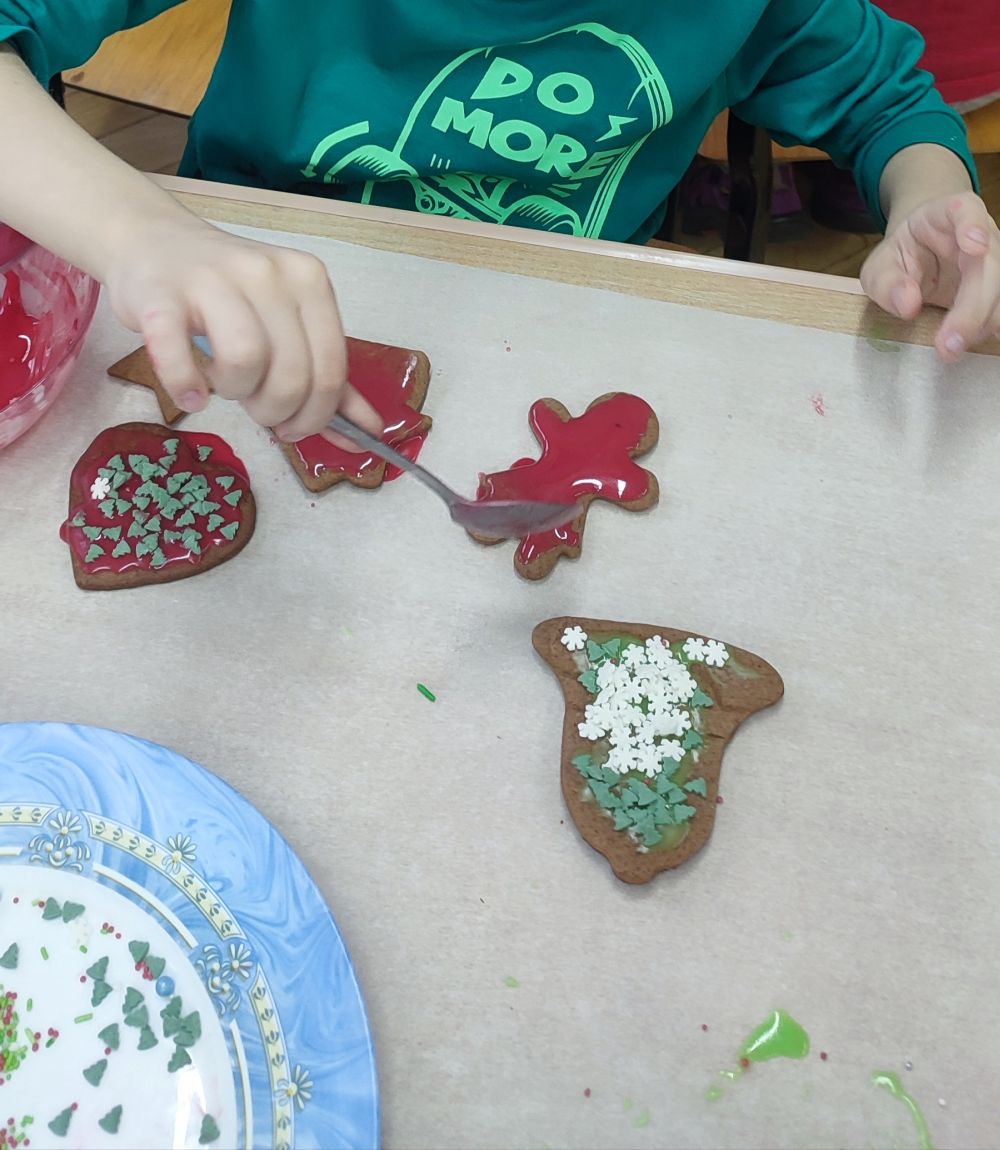 Na zdjęciu widać pierniczki leżące na stole, które dziecko ozdabia czerwonym lukrem.