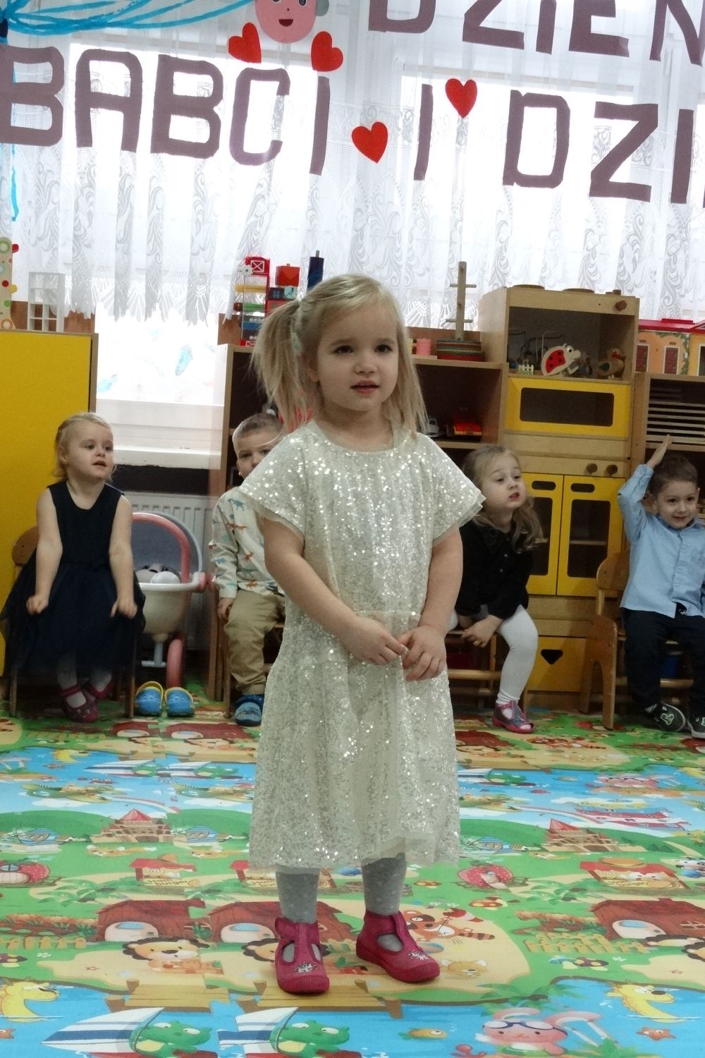 Dziewczynka w białej sukience stoi na środku sali i recytuje wierszyk