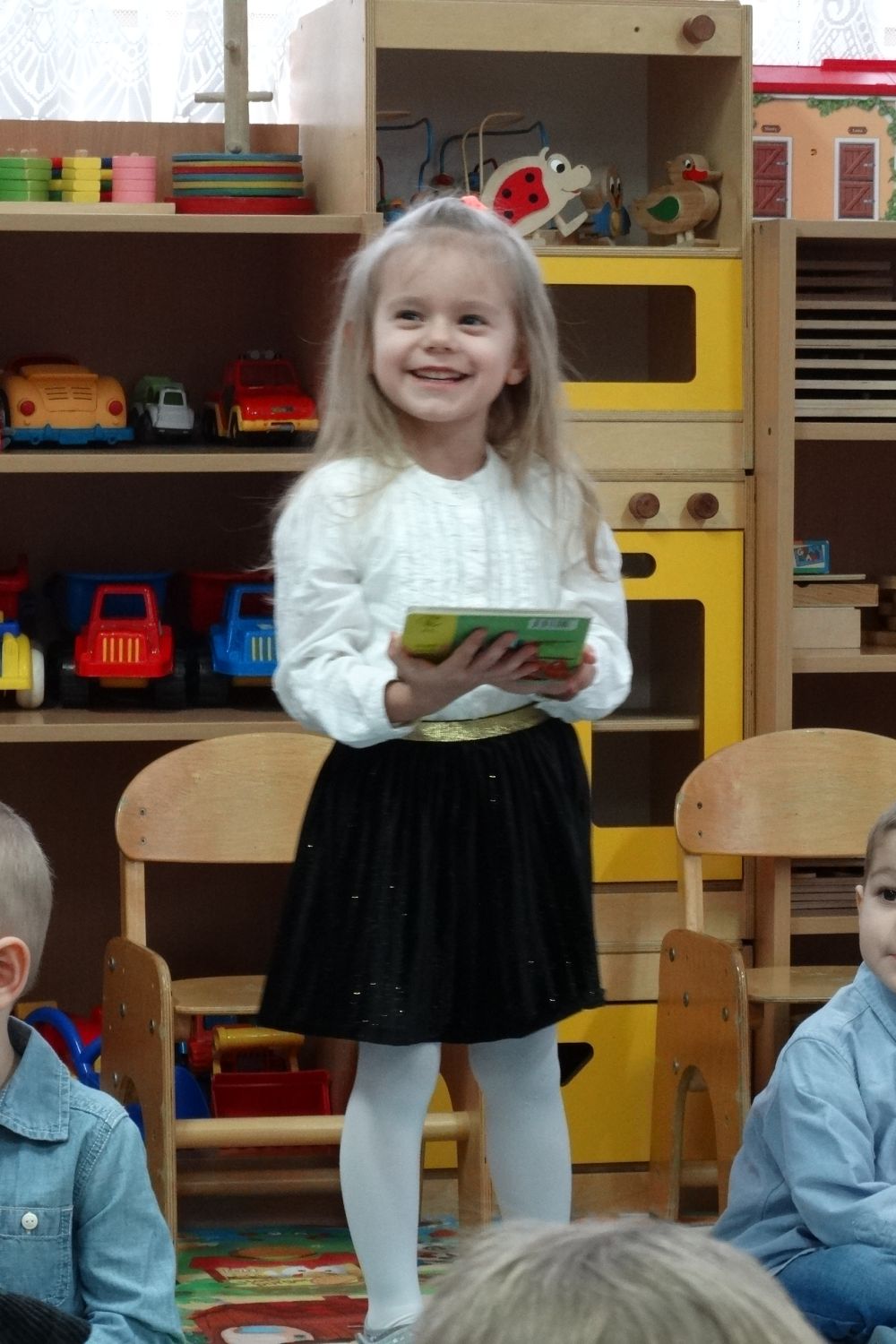 Uśmiechnięta dziewczynka w białej bluzce i czarnej spódniczce stoi na środku sali i trzyma w ręce książkę. Z tyłu siedzą inne  dzieci z grupy