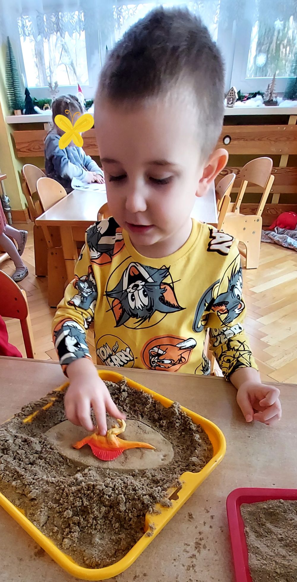 Chłopiec siedzi przy stoliku, przed nim leży miska z piaskiem w której  chłopiec trzyma dinozaura ręka