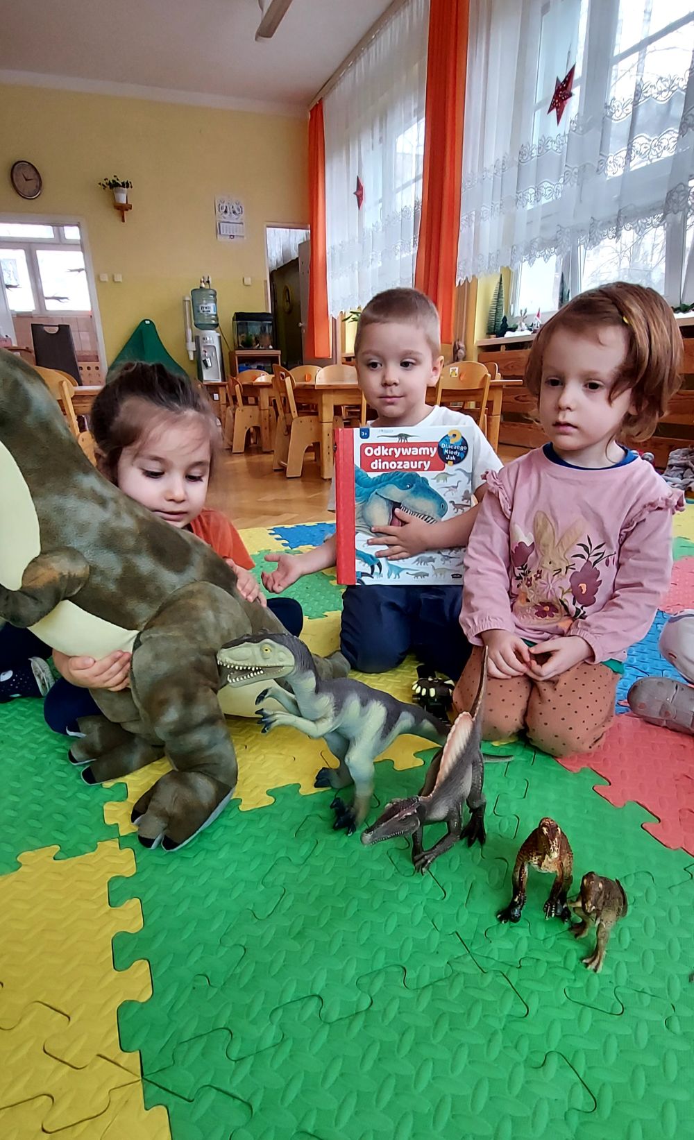 Dzieci siedzą na dywanie a przed nimi stoją zabawkowe dinozaury.