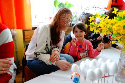 Dziewczynka pokazuje pomalowane jajko wielkanocne. Obok siedzi mama