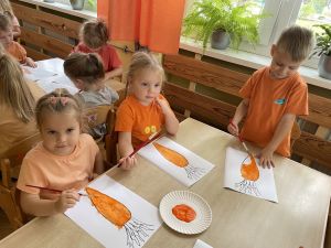 Dzieci siedzą przy stoliku i malują farbami obrazek marchewki