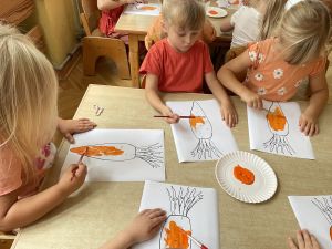 Dzieci siedzą przy stoliku i malują farbami obrazek marchewki