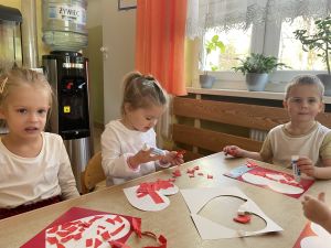 Dzieci siedzą przy stolikach i wykonują pracę plastyczną. Wyklejają serduszka czerwoną bibułą