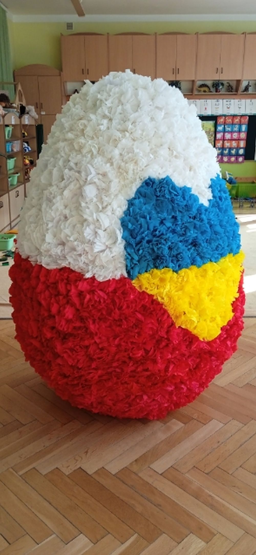 Na zdjęciu jest wielkie jajko styropianowe ozdobione różyczkami z krepiny. Jajko jest w barwach biało czerwonych, a na środku jest żółto niebieskie serce.