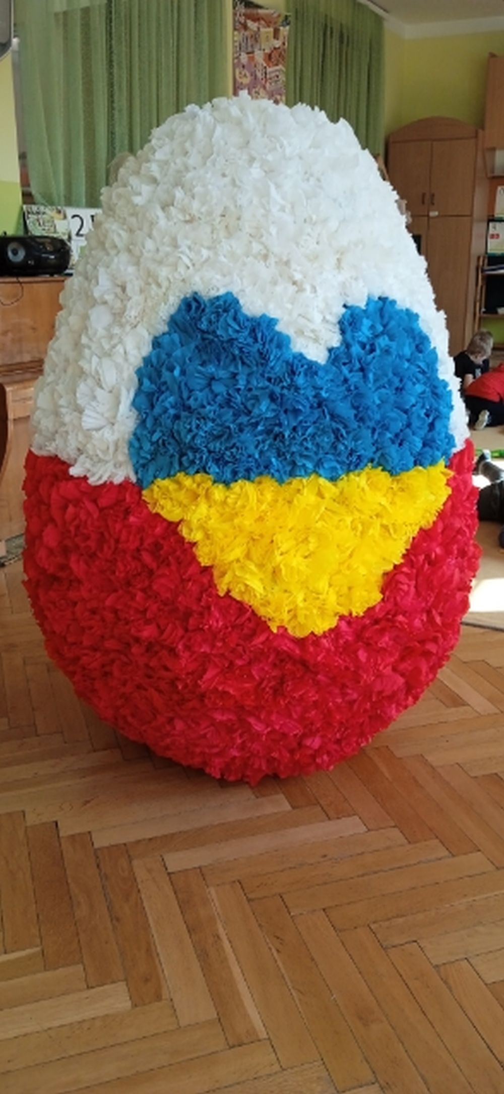 Na zdjęciu jest wielkie jajko styropianowe ozdobione różyczkami z krepiny. Jajko jest w barwach biało czerwonych, a na środku jest żółto niebieskie serce.