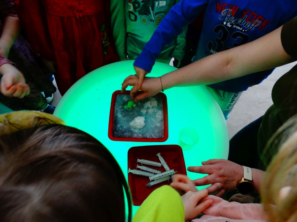 Dzieci stoją przy stoliku świetlnym a pani wyjmuje dinozaura z tacki z rozpuszczonym musującym jajem
