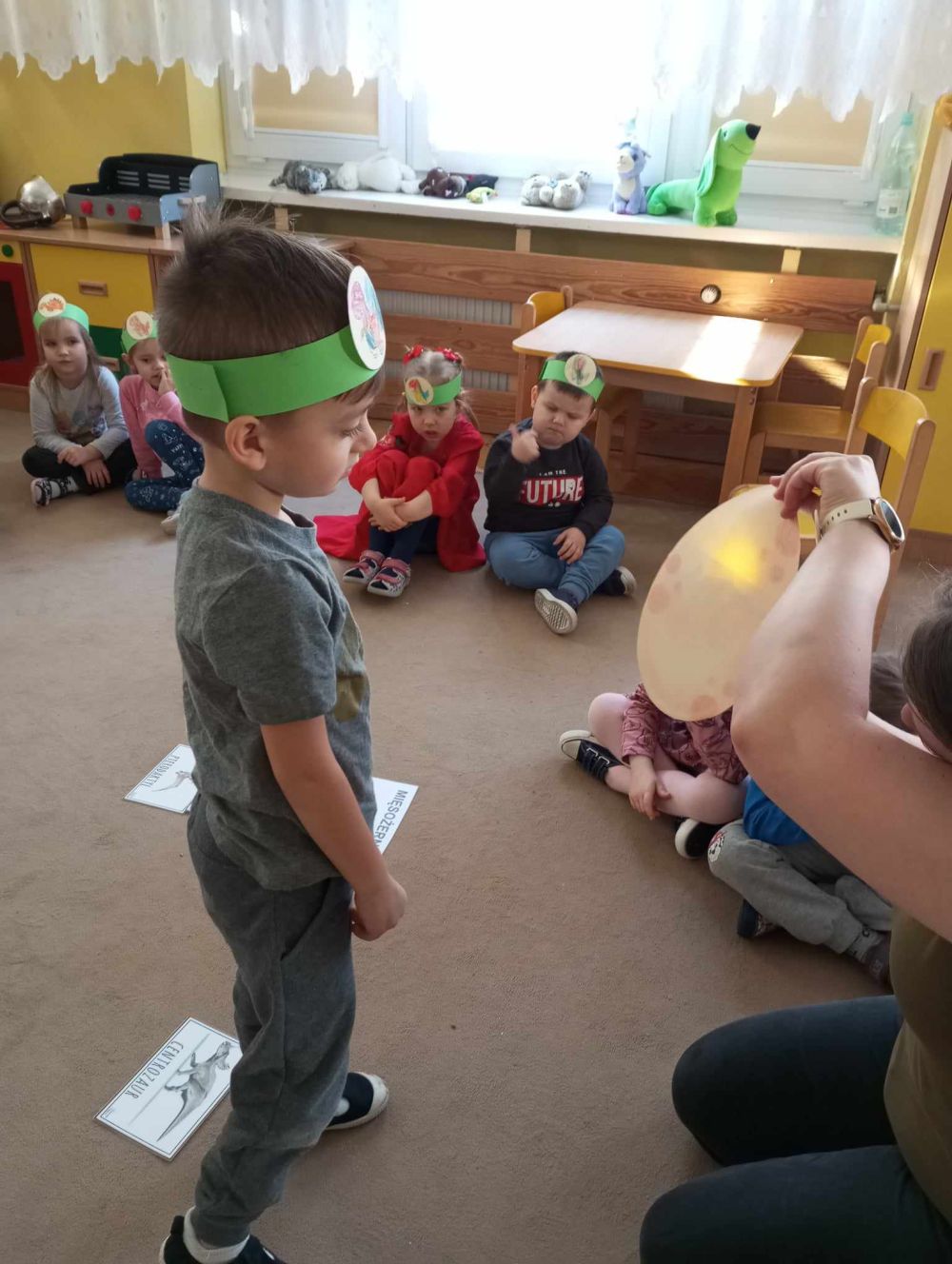 Chłopiec patrzy na papierowe jajko, dzieci siedzą na dywanie. na głowie mają opaski z obrazkiem dinozaura