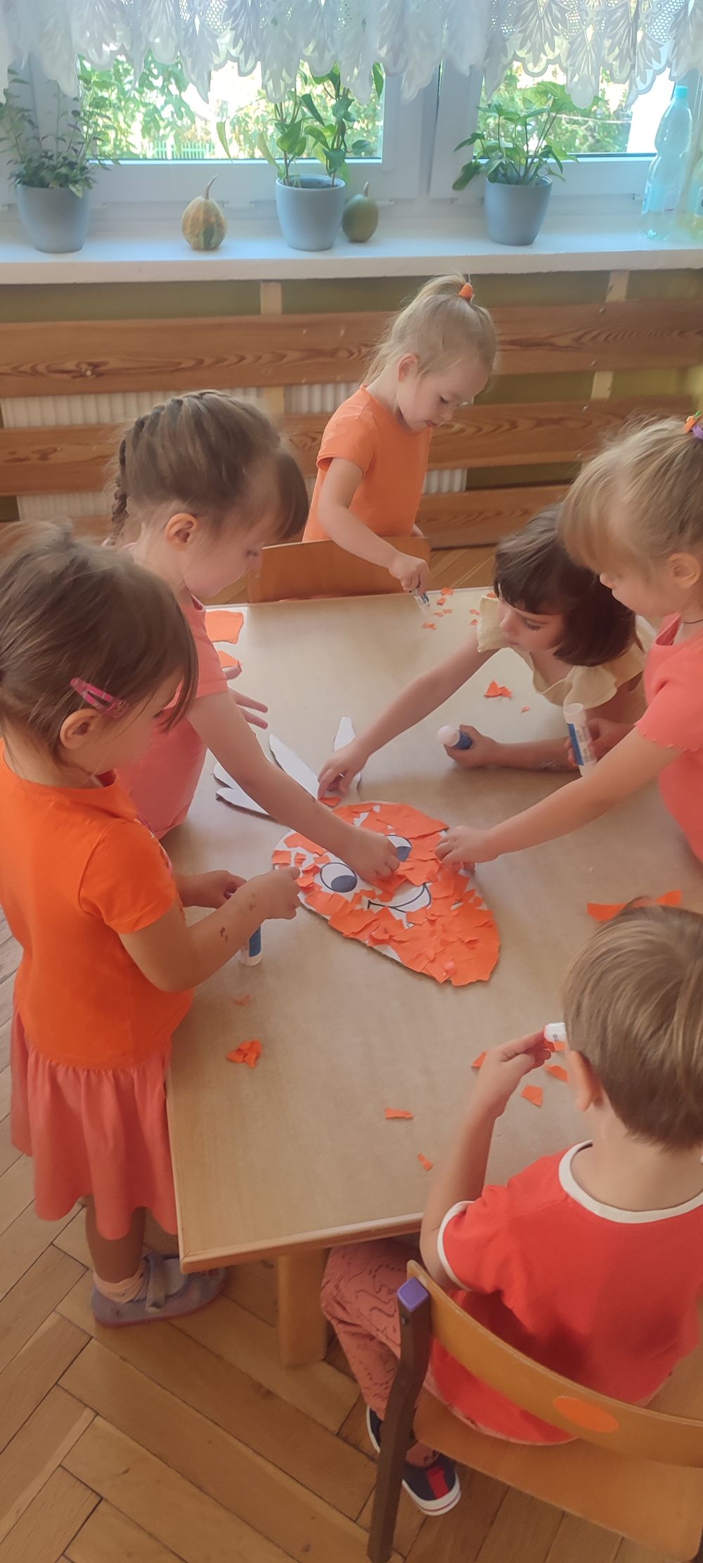 Dzieci stoją przy stoliku i wykonują pracę plastyczną. Wyklejają pomarańczowymi kartkami papieru dużą marchewkę z kartonu