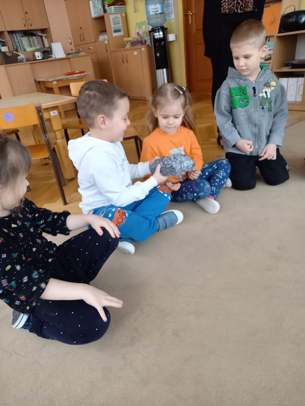 Dzieci siedzą na dywanie i podają sobie krzem