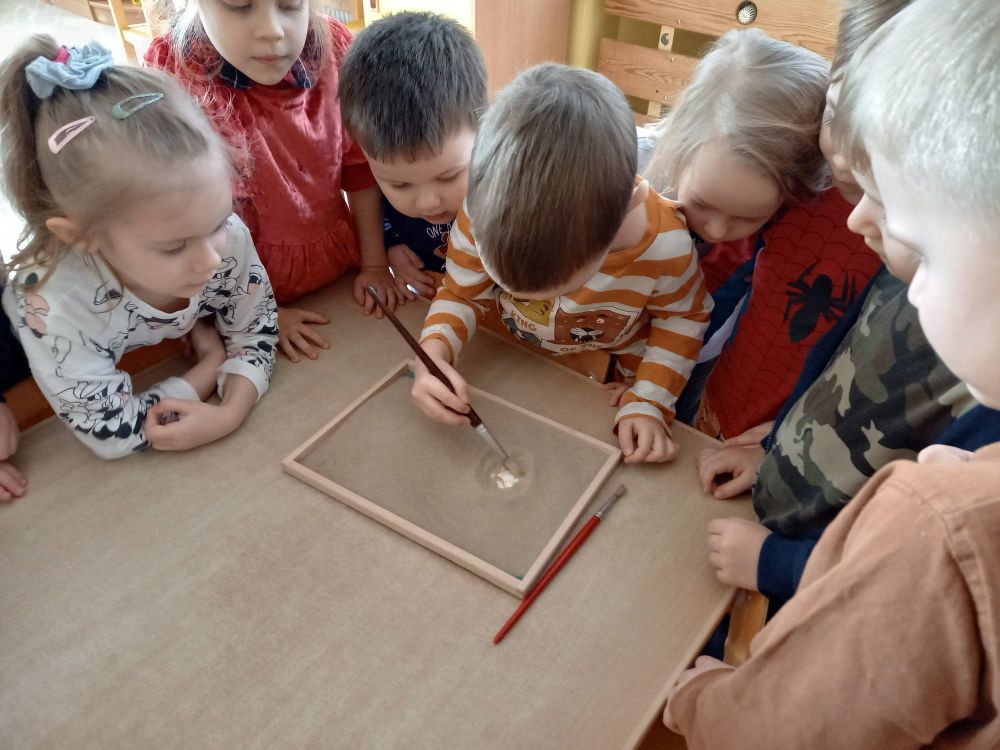 Dzieci stoją przy stoliku i pędzelkiem odkrywają piasek pod którym są gipsowe odlewy odcisków łap dinozaura