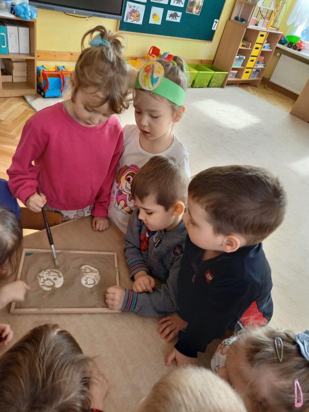 Dzieci stoją przy stoliku i pędzelkiem odkrywają piasek pod którym są gipsowe odlewy odcisków łap dinozaura