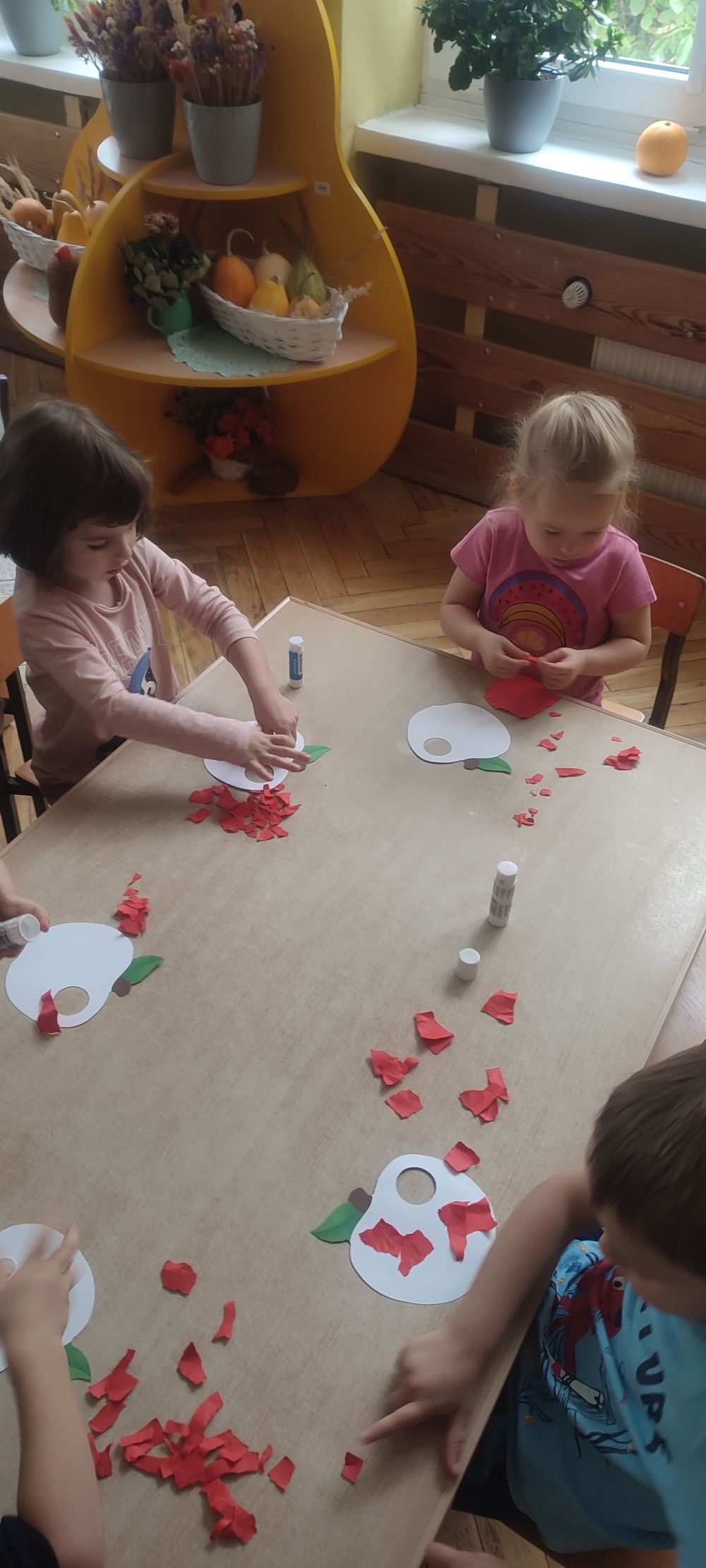 Dzieci siedzą przy stole i wyklejają jabłka czerwonymi kawałkami kartki papieru