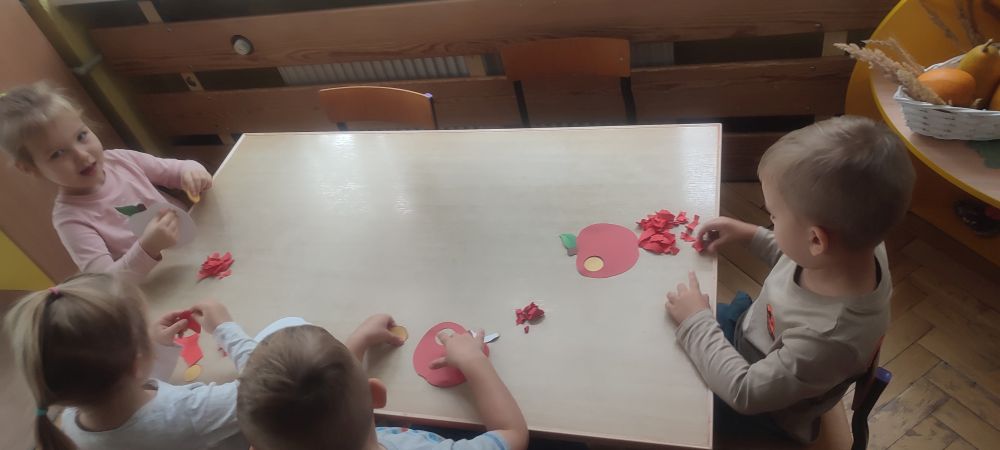 Dzieci siedzą przy stole i wyklejają jabłka czerwonymi kawałkami kartki papieru