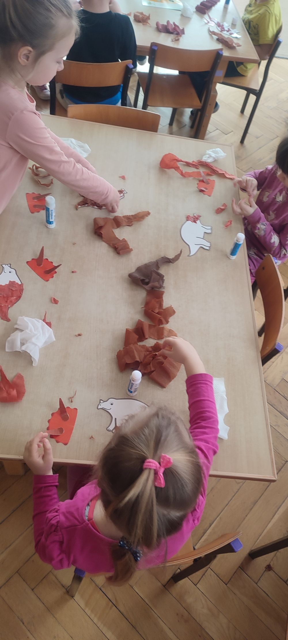 Dzieci siedzą przy stolikach i wyklejają obrazek dinozaura bibułą