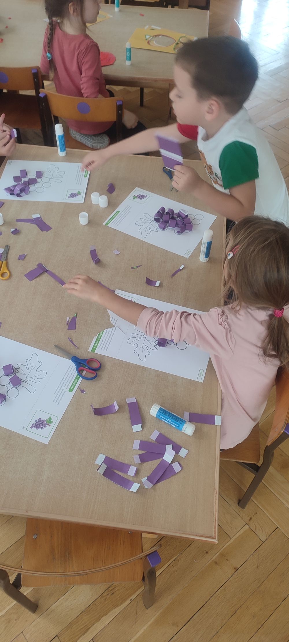 Dzieci siedzą przy stole i wyklejają paskami papieru winogrona