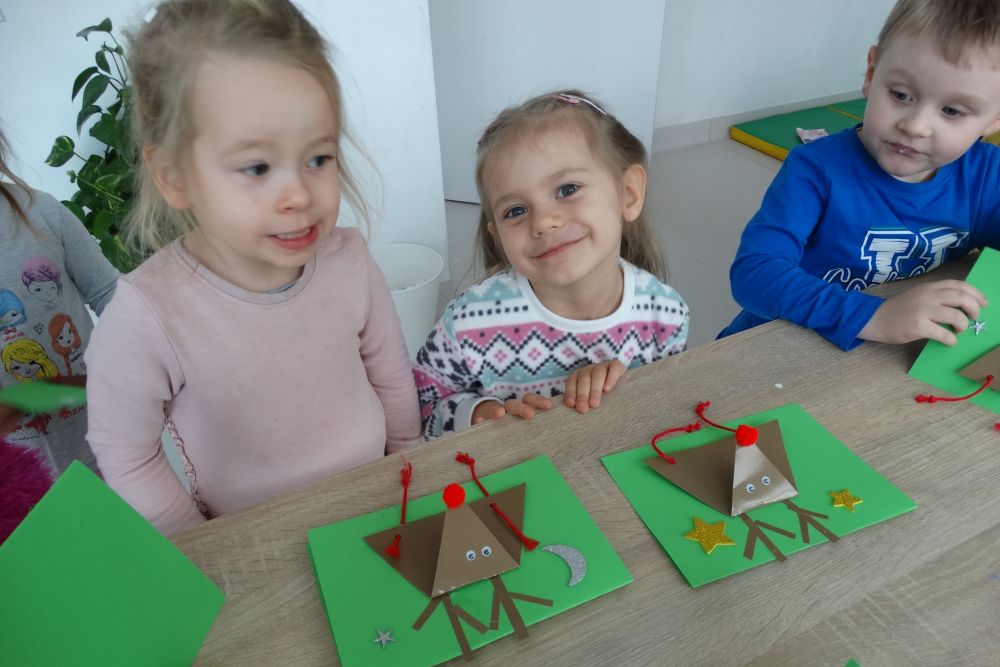 dwie dziewczynki i  jeden chłopiec stoją przy stoliku i pokazują zrobione przez siebie kartki 
