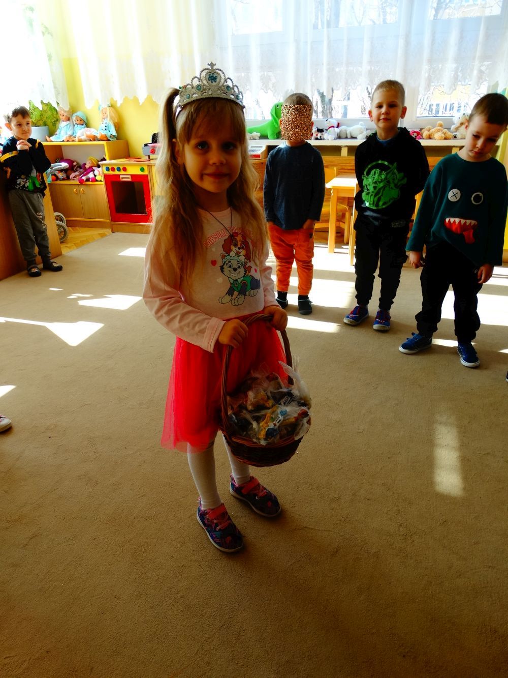 Dziewczynka z diademem na głowie stoi na środku sali i w koszyczku ma cukierki dla dzieci. Z tyłu stoją chłopcy