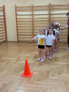 Dziewczynki stoją na sali gimnastycznej w rzędzie a przed nimi stoi pachołek