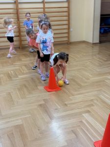 Dziewczynki stoją na sali gimnastycznej w rzędzie a przed nimi stoi pachołek. Jedna dziewczynka toczy piłkę