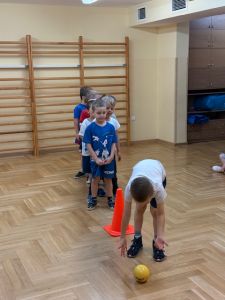 Chłopcy stoją na sali gimnastycznej w rzędzie a przed nimi stoi pachołek. Jeden chłopiec toczy piłkę