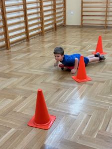 Chłopiec jedzie slalomem między pachołkami na deskorolce gimnastycznej leżąc na brzuchu