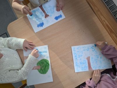 Dziecko maluje tło na kartce z drzewem i chmurami niebieską bibułą