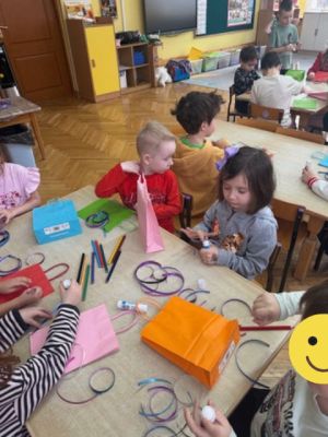 Dzieci siedzą przy stolikach i ozdabiają papierowe torebki