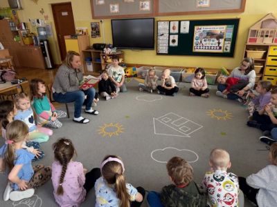 Mama Wiktorii czyta dzieciom książkę. Dzieci siedzą w kole na dywanie