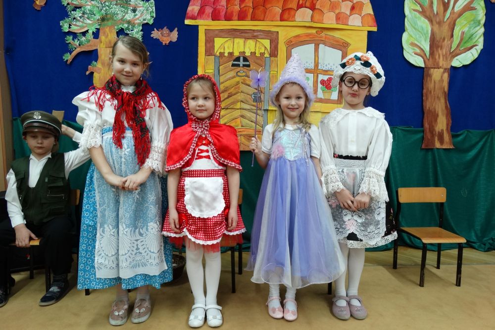4 dziewczynki stoją na tle chatki i drzew zrobionych z papieru. Zaczynając od lewej: dziewczynka w stroju mamy, dziewczynka w stroju czerwonego kapturka, dziewczynka w stroju motylka, dziewczynka w stroju babci