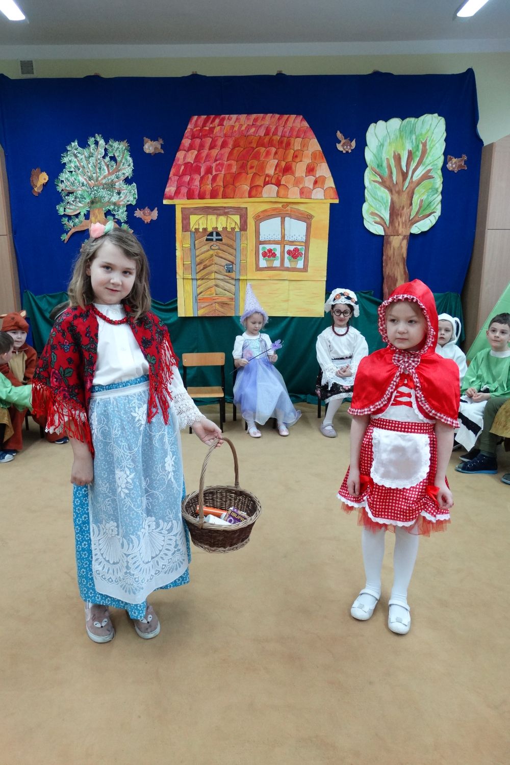 Na zdjęciu widać dwie dziewczynki w stroju mamy i czerwonego kapturka, które stoją na środku sali. Za nimi siedzą inne dzieci z grupy