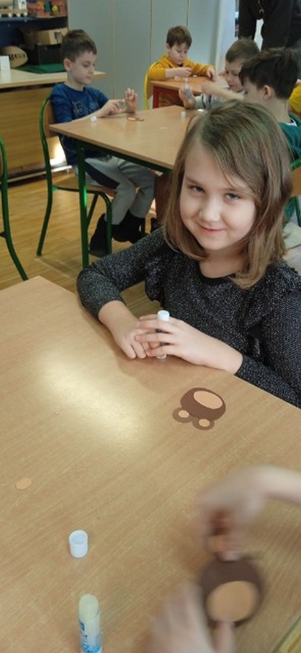 Na pierwszym planie widać dziewczynkę siedzącą przy stoliku na którym leży papierowy miś, w tle inne dzieci siedzące przy stolikach robiące papierowe misie