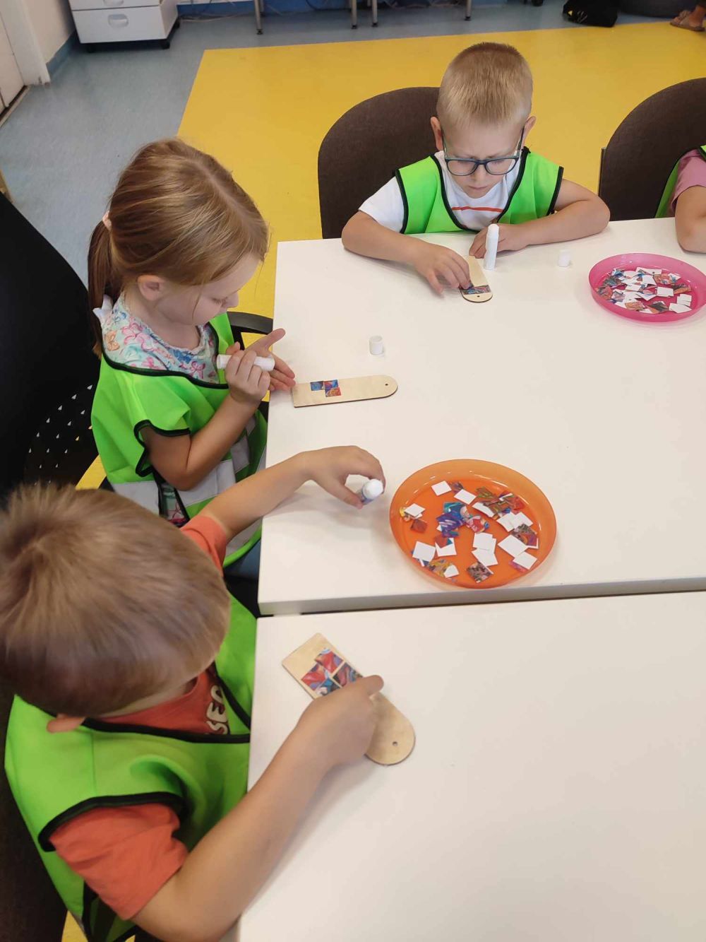 Dzieci c grupy 4 siedzą przy stołach w bibliotece i naklejają kolorowe karteczki na drewniane zakładki do książek