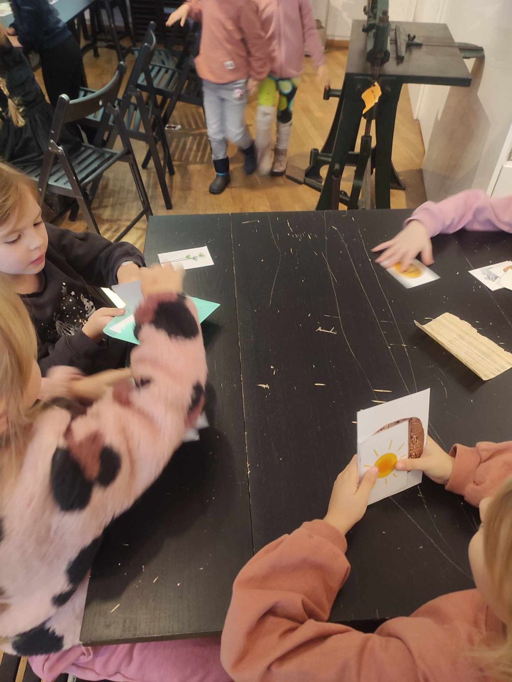 Dzieci siedzą przy stoliku w muzeum i trzymają w rękach karteczki z kolorowymi obrazkami