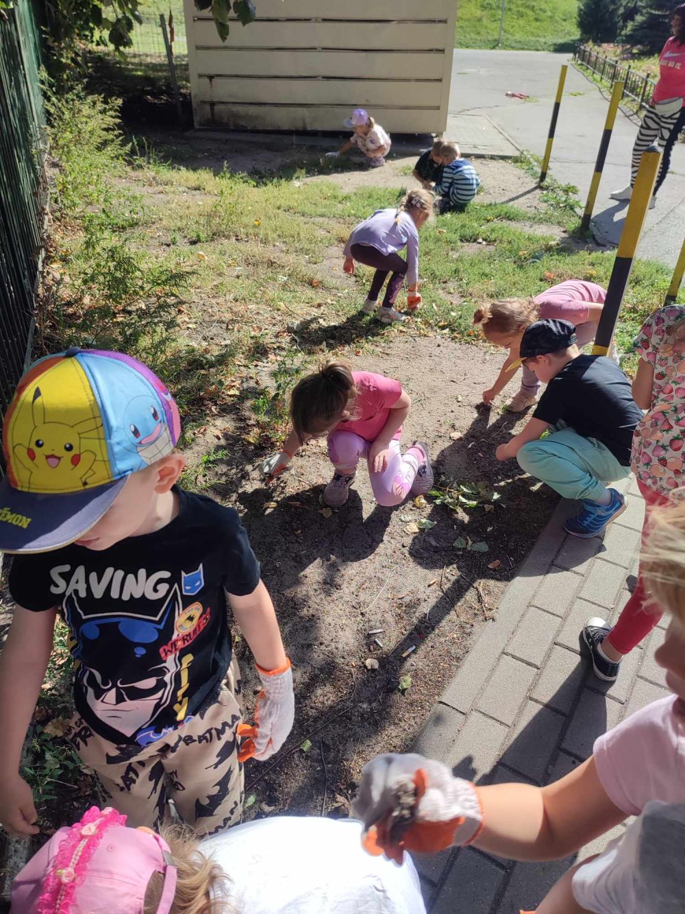 Dzieci zbierają śmieci z trawnika przy płocie przedszkola
