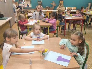Dzieci siedzą przy stolikach. Przed nimi leży niebieska kartka z przyklejoną fioletową kartką w kształcie doniczki.