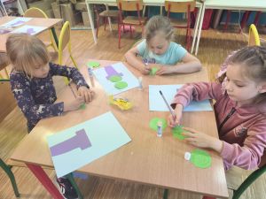 3 Dziewczynki siedzą przy stoliku. Przed nimi leży niebieska kartka z przyklejoną fioletową kartką w kształcie doniczki. Na zielonych kółkach z papieru dziewczynki rysują kolce ołówkiem