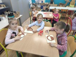 Na zdjęciu na pierwszym planie widać 3 dziewczynki siedzące przy stole. Na stole leżą kartki  dla mam, które robią dzieci. Na drugim planie 3 dzieci siedząca przy stole
