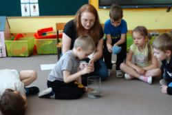 Dzieci siedzą na dywanie a jeden chłopiec wkłada coś do pojemnika z wodą
