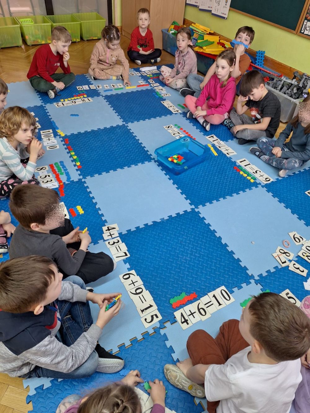 Dzieci siedzą na dywanie a przed nimi leżą karteczki z działaniami i kolorowe klocki
