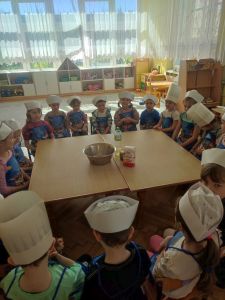 Dzieci ubrane w fartuszki i czepki kucharskie siedzą przy stole na którym stoją miska i produkty do zrobienia chlebka