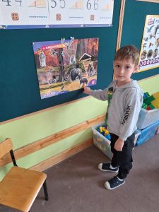 Chłopiec stoi i pokazuje palcem dzika na plakacie przedstawiającym las.