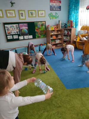 Dzieci stoją na dywanie i wykonują skłon trzymając butelkę w rękach