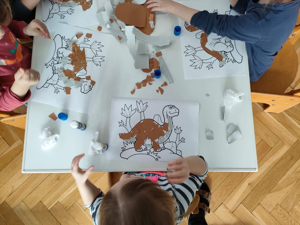 Dzieci siedzą przy stoliku i na obrazek z dinozaurem naklejają kawałki kartki