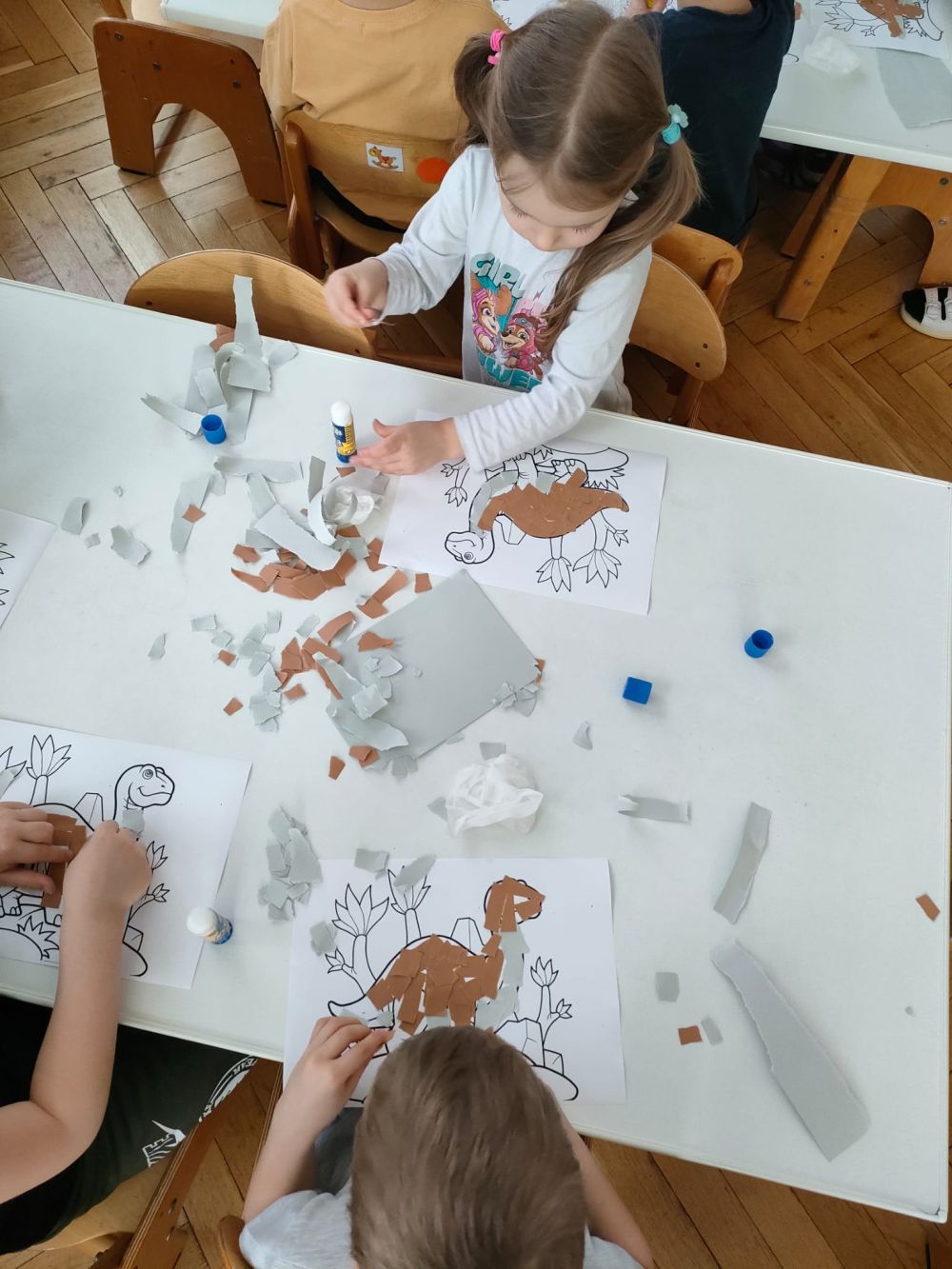 Dzieci siedzą przy stoliku i na obrazek z dinozaurem naklejają kawałki kartki