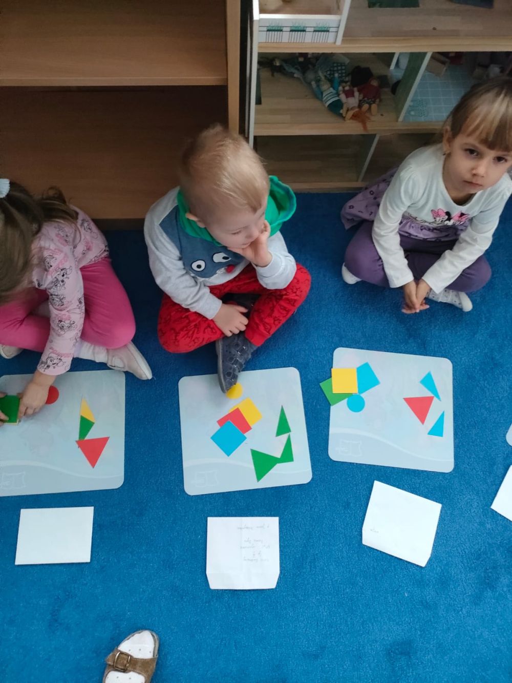 Dzieci siedzą na dywanie a przed nimi na białych podkładkach leżą papierowe figury geometryczne