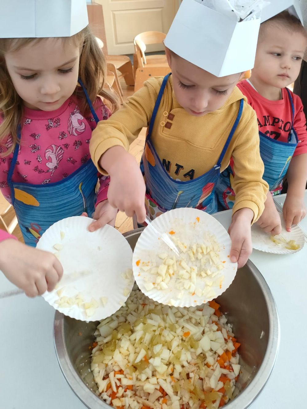 Chłopiec i dziewczynka w strojach kucharzy zrzucają z talerzyków pokrojone warzywa
