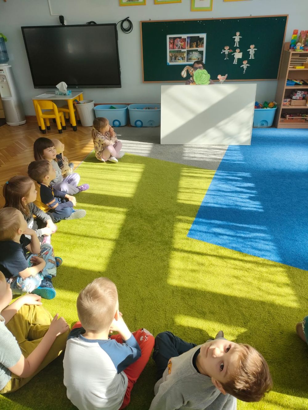 Dzieci siedzą na dywanie i oglądają przedstawienie kukiełkowe robione przez nauczycielkę