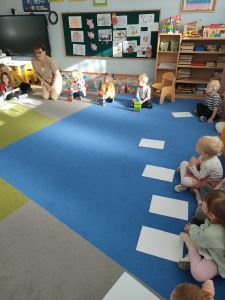 Na dywanie siedzi nauczycielka i dzieci. Troje dzieci buduje z klocków domki